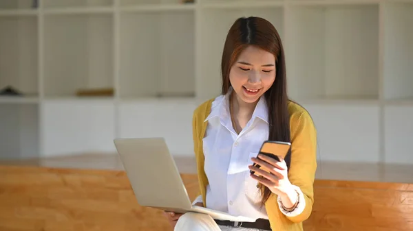 Photo of beautiful creative woman putting laptop on her legs and holding smartphone in hand white sitting at the modern wooden bench. Comfortable cafe/common area concept.