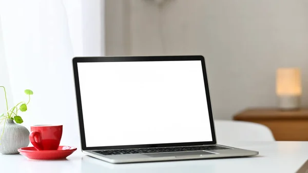 Laptop computer white screen putting together with potted plant and coffee cup on the modern table with comfortable living room as background.