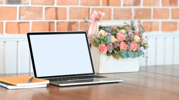 Photo of white blank screen laptop, notebook, pen and roses bouquet in the white plastic basket putting on the wooden table with modern concrete wall as background.