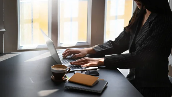 Cropped shot executive woman in black striped suit typing on computer laptop keyboard while sitting at the modern working table with office windows as background.