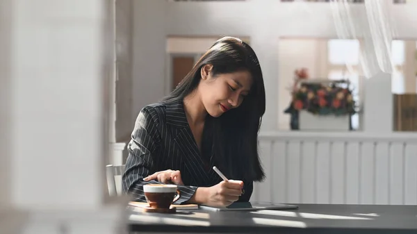 Photo of young executive woman in black striped suit drawing business plan by computer tablet at the modern working desk with comfortable office as background.