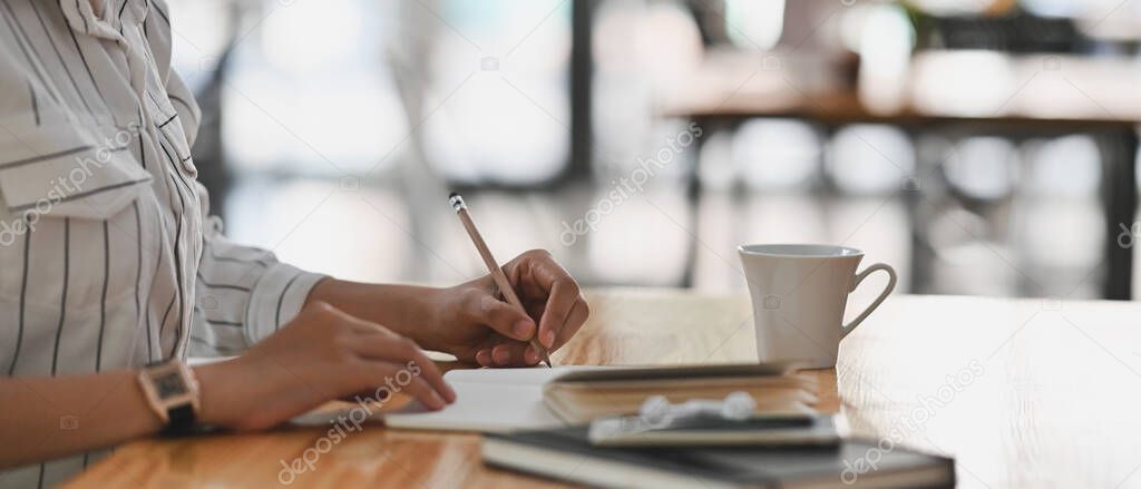 Cropped shot of young secretary woman in white striped shirt sitting at the wooden desk and writing on the notebook/taking notes.