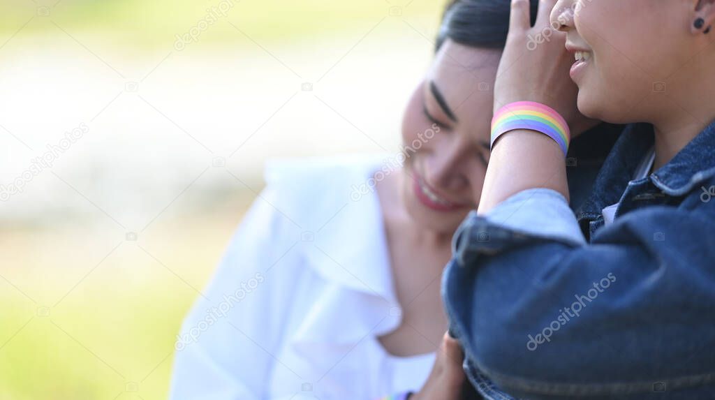 Photo of young LGBT lesbian couple embracing while sitting together at the grass field with blurred sunny outdoors/park as background. LGBT happiness concept.