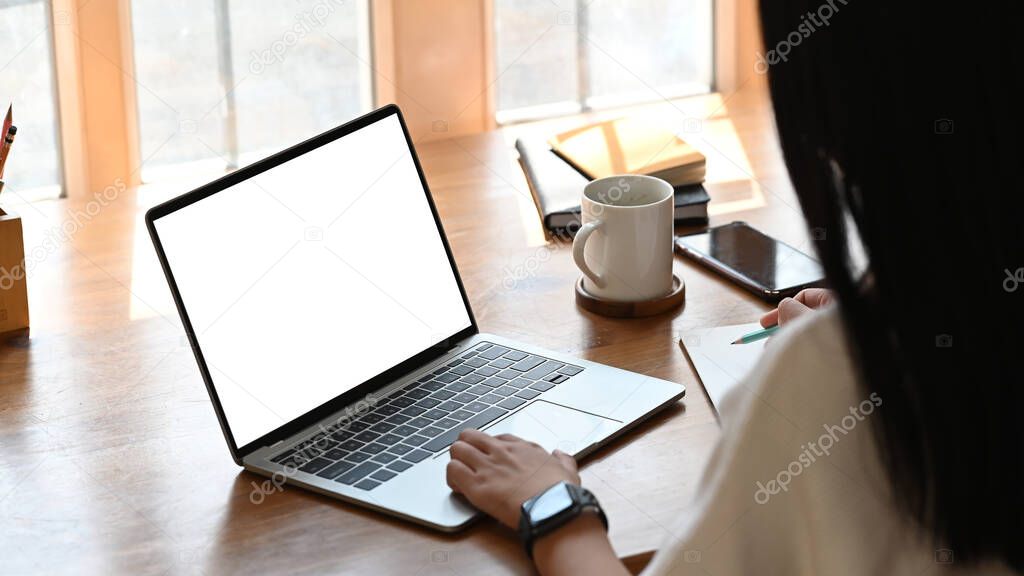 Cropped shot of young designer woman writing on notebook while she sitting and using white blank screen laptop at the modern wooden table. Women lifestyle concept.
