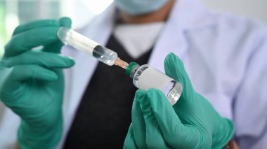 Doktorun eli yakından görülüyor. Elinde şırınga olan tıbbi eldiven var. Şırınga aşısını şişeden alıyor..