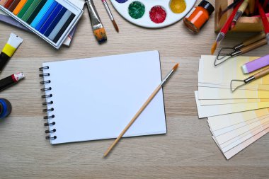Renk örnekleri, boya fırçaları ve ahşap masadaki diğer çeşitli ekipmanlarla çevrili defterli sanatçı işyeri.