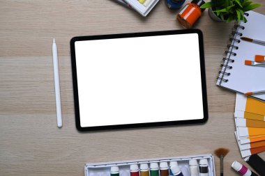 Sanatçı veya tasarımcı çalışma alanının dijital tablet, stilus kalem ve tahta masadaki boyama araçlarıyla üst görünümü.