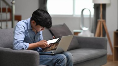 Genç Asyalı serbest çalışan, dizüstü bilgisayarda çalışan, dijital tablet kullanan ve evdeki kanepede oturan.