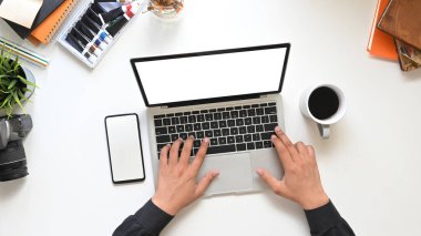 Üst görüşlü yaratıcı adamın elleri bilgisayar laptopunda beyaz beyaz ekranlı kahve fincanı, boş ekran akıllı telefon ve aksesuarlarla çevrili çalışma masasına koyuyor..