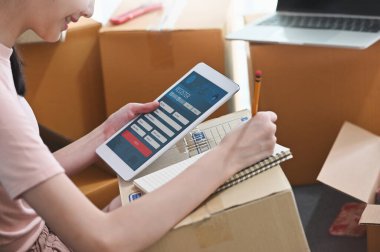 Genç ve güzel bir kadının eşyalarını müşteriye teslim etmek için konforlu oturma odasında bilgisayar tableti ve dizüstü bilgisayar kullanarak paketlediği fotoğraf. Çalıştır / Girişimci konseptini.