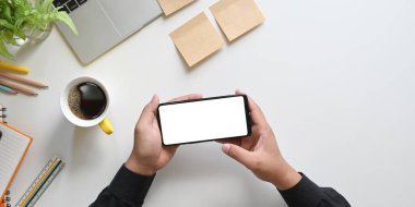 Üst görüntüde bilgisayar, kahve fincanı, kalemler, günlük, not defteri ve saksı bitkisi, cetvel ile birlikte beyaz ekranda beyaz ekranlı siyah bir akıllı telefonu tutan eller görülüyor..