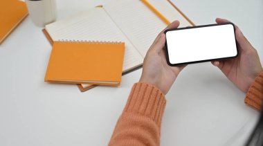 Arkaplan olarak çalışma masasının üzerinde beyaz boş ekran ile kırpılmış siyah bir akıllı telefon tutan ellerin kesilen görüntüsü.