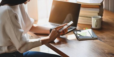 Bilgisayarın önündeki ahşap masada otururken elinde akıllı telefon tutan güzel bir kadının fotoğrafı. Laptop, kahve fincanı, kitap yığını ve düzenli bir kütüphanede defter..