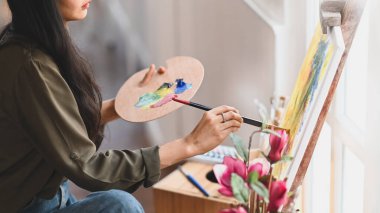 Çağdaş sanat stüdyosunda otururken elinde fırça tutan ve tuvale yağlı boya renkleri çizen genç bir kızın kırpılmış görüntüsü. Sanatçı olarak yaratıcı kadın kavramı.
