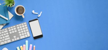 Üzerinde aksesuarlar olan çalışma masasının üst görünümü. Beyaz beyaz ekran akıllı telefon, kahve fincanı, kablosuz kulaklık, defter, kalem ve kablosuz klavye mavi boşlukta yatıyordu..