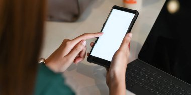 Arkaplan olarak çalışma masasının üzerinde beyaz boş ekran ile kırpılmış siyah bir akıllı telefon tutan ellerin kesilen görüntüsü.