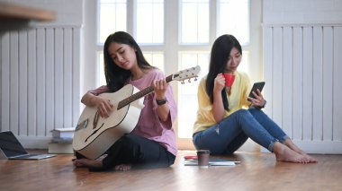 Arka plan olarak rahat oturma odası / müzik stüdyosu üzerinde ahşap zemin katta birlikte otururken akustik gitar çalmayı öğreten / öğrenen genç kadınların fotoğrafı.