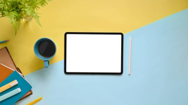 Çalışma alanının düz beyaz ekran bilgisayar tableti, kahve fincanı, stilus kalem, işaret kalemleri, renkli çalışma masasına dizilmiş defter ve saksı bitkisi ile üst görünümü.