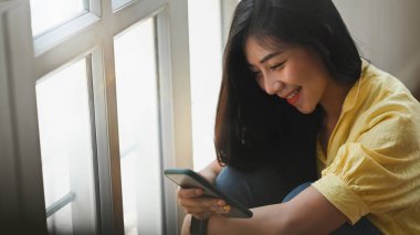 Otururken akıllı telefon kullanan güzel bir kadın oturma odasının arka planında dizlerine sarılıyor..