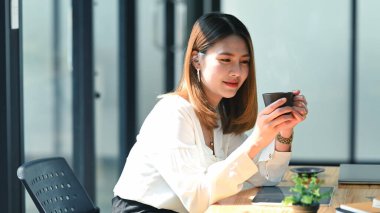 İş kadını elinde bir kahve fincanı tutarken, modern ofis arka planında, ahşap bir çalışma masasının önünde oturuyor..