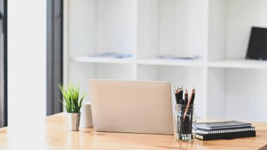 Bilgisayar dizüstü bilgisayarı, kalem tutacağı, saksı bitkisi, defter ve kahve fincanı arka plan olarak ofis kitaplığı rafı ile modern ahşap masaya konuyor..