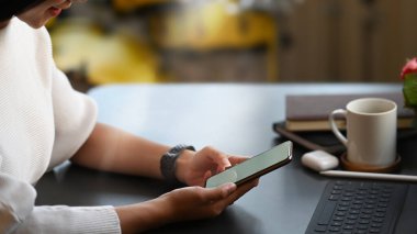 Genç yaratıcı kadının elleri boş ekranlı bir akıllı telefonu tutarken rahat oturma odasının üstünde arka plan olarak otururken görüntüsü kesilmiş..