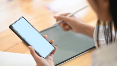 Modern ofisteki ahşap çalışma masasında beyaz ekranlı tablet kullanırken ve otururken elinde akıllı telefon ve stilus kalem tutan sekreter kadının kesilmiş görüntüsü..