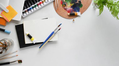 Çalışma masasında çalışan ressamın üst görünümü. Renkli tüp, saksı bitkisi, boya fırçası, durağan, renk paleti ve not defteri düz beyaz izole arkaplan üzerinde yatıyordu. Dağınık çalışma alanı kavramı.