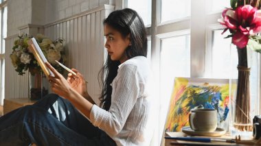 Resim, elinde boya fırçası olan genç ve güzel bir sanatçının resim yaparken sanat aksesuarlarının yanına oturması ve arka plan olarak stüdyo pencereleri olan küçük ahşap bir masa koyması..