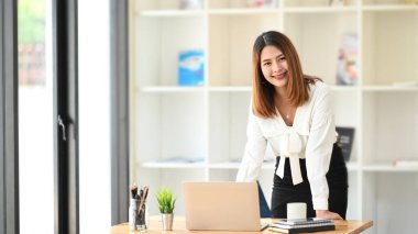 Sekreter olarak çalışan güzel bir kadının fotoğrafı gülümseyen ve ahşap çalışma masasında duran rahat ofis kitaplığı arka planda..