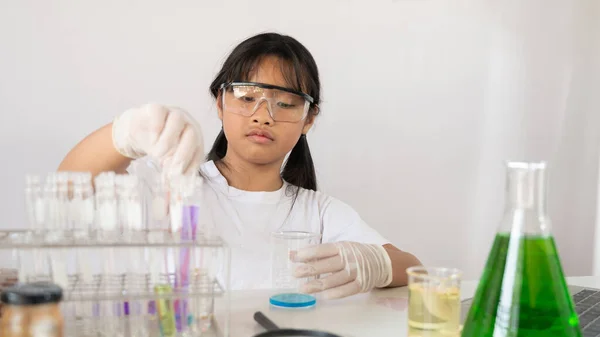 科学実験をしながら化学ガラスを持っている若い可愛い女の子の写真と背景として白い実験室の壁と現代の白いテーブルに座って — ストック写真
