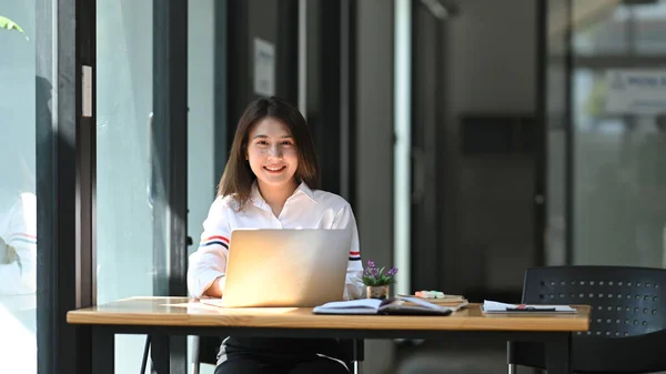 照片上 一个漂亮的女孩笑着坐在她的笔记本电脑前 坐在舒适客厅上方的木制工作台前作为背景 — 图库照片