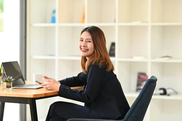 スマートフォンを持っている魅力的なビジネス女性の写真と背景として快適なオフィスのガラスの壁とモダンな木製のテーブルに座って笑顔 — ストック写真