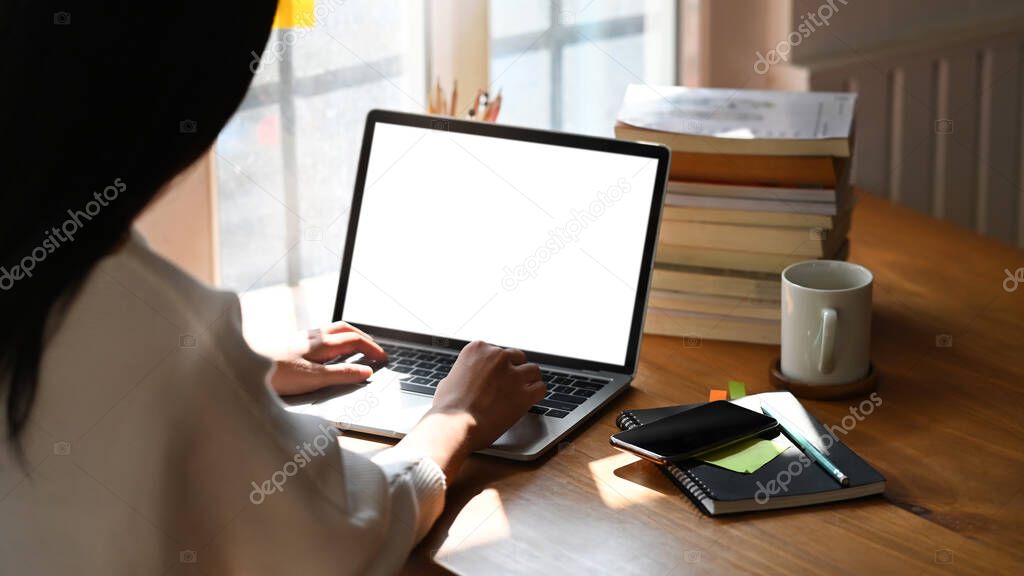 Cropped image of secretary woman's hands typing on computer laptop with white blank screen that putting on orderly working desk that surrounded with notes, coffee cup and stack of books.
