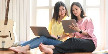 Arka plan olarak rahat müzik stüdyosu pencereleri üzerinde ahşap zeminde otururken ellerinde bilgisayar tableti ve laptopuna bakan kız kardeşler..