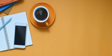 Siyah boş ekranlı kesilmiş beyaz akıllı telefonun üst görüntüsü kahve fincanı, not, kalem, saksı bitkisi ve defterle renkli çalışma masasına konuyor. Renkli çalışma alanı kavramı.