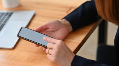 Arka plan olarak rahat oturma odasının üstündeki ahşap çalışma masasında otururken ve dinlenirken, yakın plan kadının elleri akıllı telefonu tutuyor ve kullanıyor..