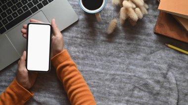 Eller 'in bilgisayar dizüstü bilgisayarı, eski kitaplar, kahve fincanı ve masa örtüsüyle kaplı çalışma masasının üzerine yerleştirilmiş yabani otları üzerinde beyaz ekran akıllı telefonu tutarken en üstteki görüntü..