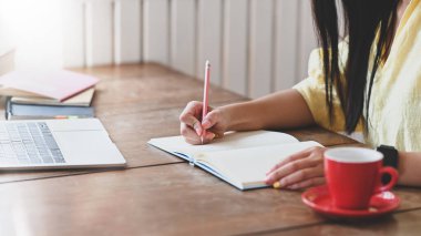 Sarı pamuklu tişörtlü güzel bir kadının bilgisayarının önünde oturmuş not alırken ahşap çalışma masasına kitap yığını ve kahve fincanı koyduğu fotoğraf..