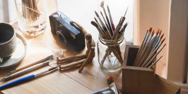 Cam vazodaki boya fırçası, ahşap çalışma masasına yerleştirilmiş. Kamera, ahşap kukla, boya fırçası ve kahve fincanı ile çevrili. Dağınık ressam çalışma masası konsepti.