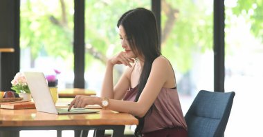 Asyalı bir kadın ahşap çalışma masasında otururken bilgisayar kullanıyor..