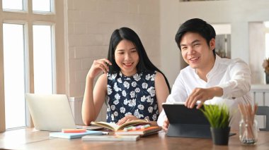 Web sitesi tasarımcısı olarak çalışan genç çift, arka plan olarak rahat oturma odasının üzerinde ahşap masadaki dizüstü bilgisayar ve tablette yan yana oturuyorlar..