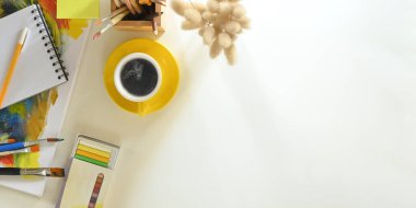 Beyaz çalışma masasının ortasına koyulan seramik kahve fincanının en üst görüntüsü ve etrafı boya fırçası, boya fırçası, kalem tutacağı, renk tüpü, not, kalem ve yabani otlarla çevrili. Ressam masasında çalışıyor..