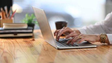 Yaratıcı bir kadının ellerinin bilgisayar laptopuna yazı yazıp ahşap çalışma masasına koyduğu ve etrafını kitap yığınları, kalem tutacağı, kahve fincanı ve saksı bitkileriyle sardığı görüntüler. Düzenli çalışma alanı.