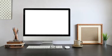 Kablosuz fare, klavye, kalem tutacağı, kitap yığını, kahve fincanı ve saksı bitkisini arka plan olarak beyaz duvarın üzerine koyarak çalışan beyaz ekranlı bilgisayar monitörü..