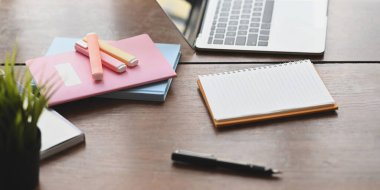 Bilgisayar dizüstü bilgisayarı ahşap çalışma masasına yerleştiriyor ve arka plan olarak defter, kalem, not, işaret kalemleri ve düzenli çalışma alanı üzerine saksı bitkileriyle çevrili..
