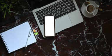 Beyaz boş akıllı telefonun, kahve fincanı, defter ve kalemle çevrili bilgisayar laptopuyla mermer dokuma masasına koyduğu en iyi görüntü. Modern ve lüks çalışma alanı kavramı.