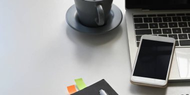 Bilgisayarın beyaz çalışma masasına koyduğu ve etrafı defterle çevrili bilgisayar görüntüsü boş ekran akıllı telefon, kahve fincanı ve kalemle çevrili. Düzenli çalışma alanı kavramı.