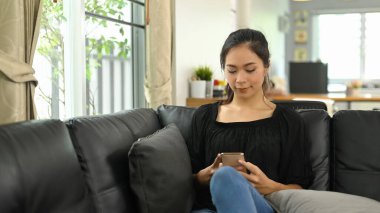 Genç Asyalı kadın siyah deri koltukta uzanırken cep telefonu kullanıyor..