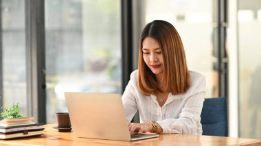 Yaratıcı bir kadının elleri bilgisayar laptopunda yazı yazarak ahşap çalışma masasına koyuyor ve etrafını kitap yığınları, kalem tutacağı, kahve fincanı ve saksı bitkileriyle çeviriyor. Düzenli çalışma alanı.